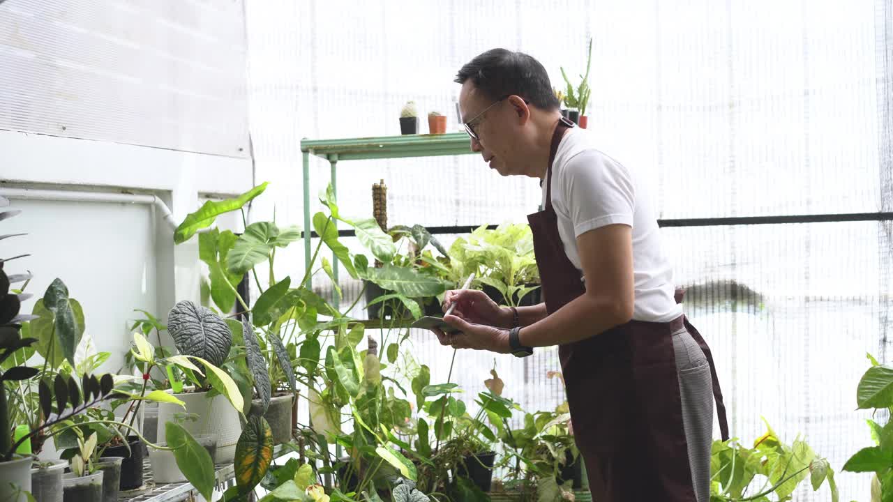 微笑的亚洲男性企业家系着围裙，用数码平板电脑检查温室里的植物。创办小型企业温室业主，中小企业，经营农业技术理念视频下载