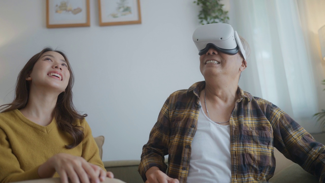 戴着虚拟现实眼镜的老爷爷和孙女坐在家里的沙发上，一起使用增强现实技术进行家庭娱乐。视频素材