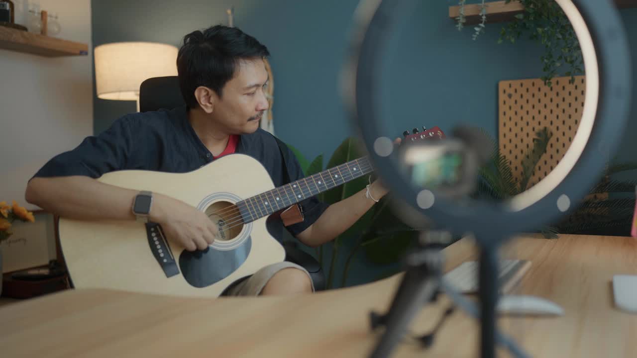 一名亚洲男子为他的博客制作视频内容，展示他的吉他技巧和唱歌。视频素材