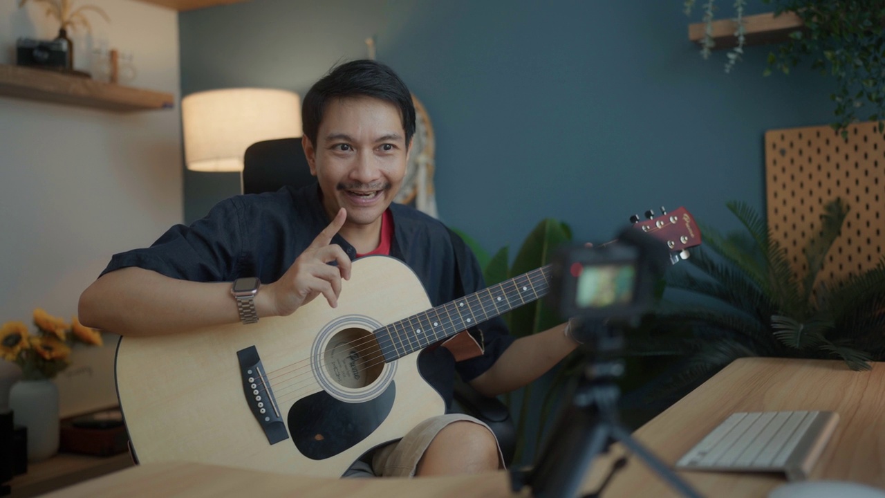 有吸引力的亚洲人有才华的艺术家录制视频的智能手机坐在桌子上在现代工作室弹吉他。视频下载
