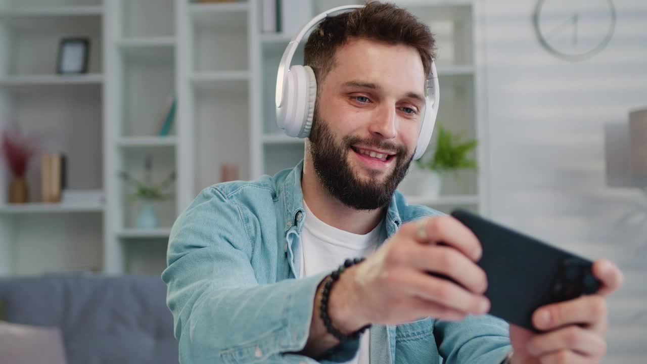 快乐的高加索人玩家戴着耳机比赛玩视频游戏在线与智能手机彩色霓虹灯光客厅现代的房子。人，电子竞技，游戏在线概念。视频下载
