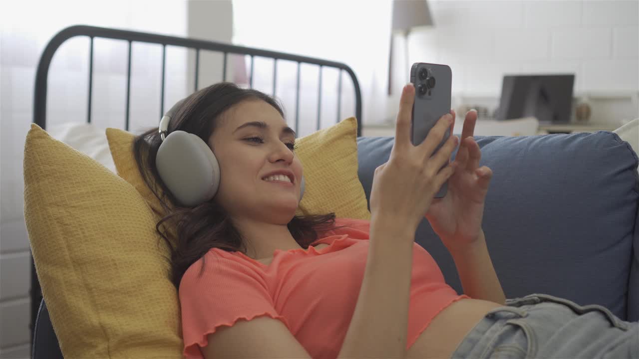 微笑的女人戴着耳机在床上玩手机视频下载