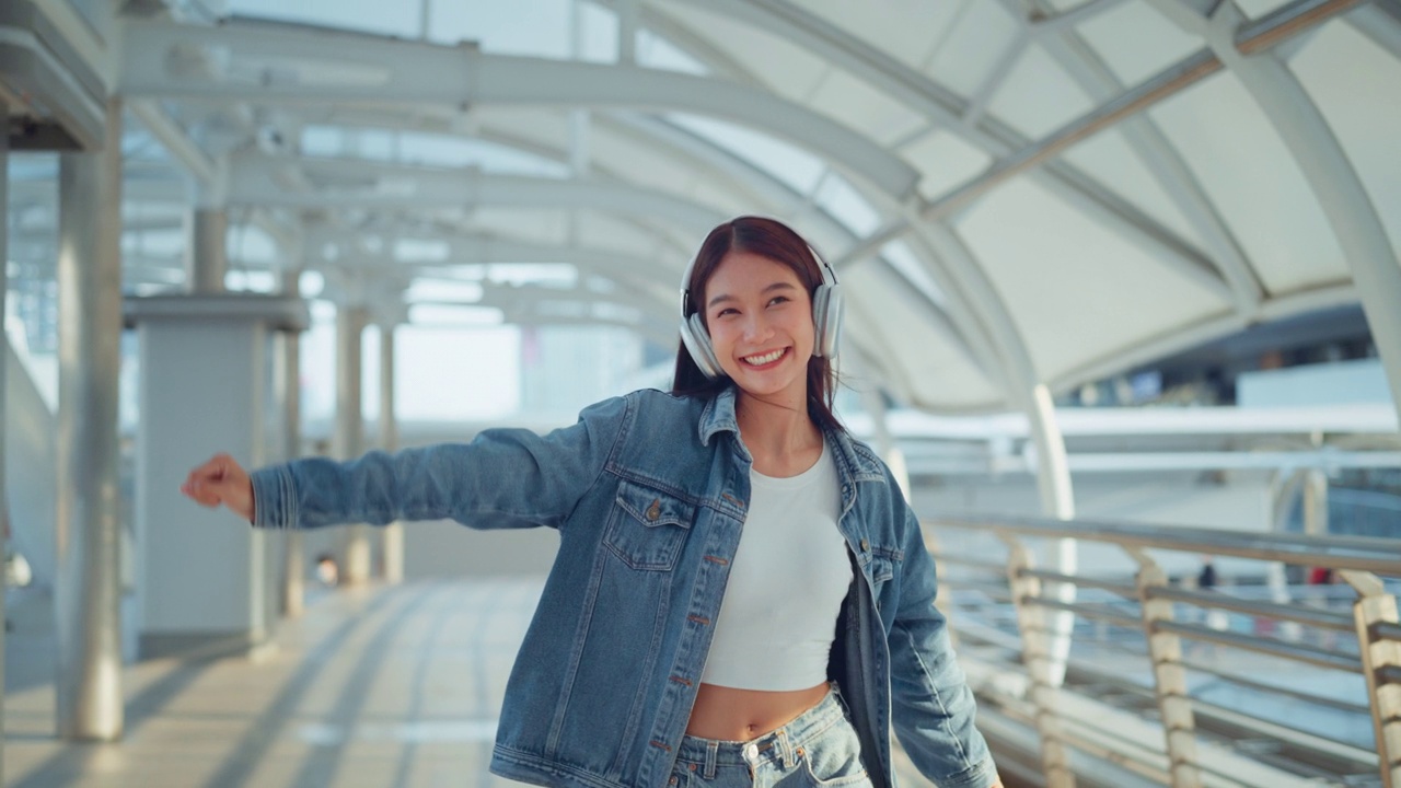 快乐的年轻亚洲女子戴着耳机在城市的街道上欣赏音乐，微笑的女性独自听音乐跳舞。视频素材