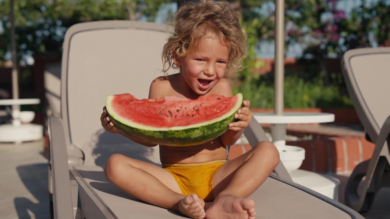 漂亮的金发男孩坐在泳池边吃西瓜，特写。快乐的2岁男孩吃着多汁的成熟西瓜。高品质4k画面视频下载