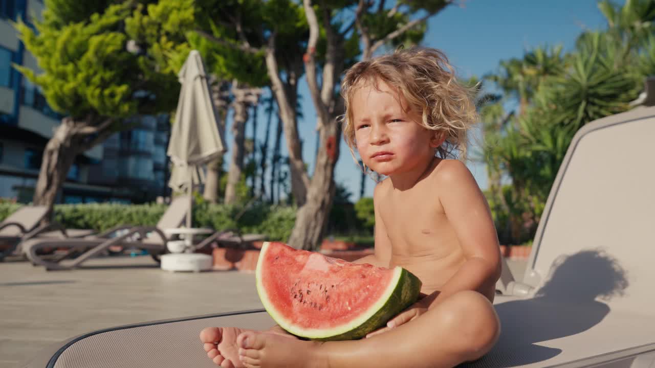 漂亮的金发男孩坐在泳池边吃西瓜，特写。快乐的2岁男孩吃着多汁的成熟西瓜。高品质4k画面视频下载
