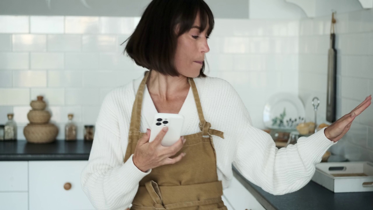 穿着围裙的无忧无虑的女人在厨房里拿着手机跳舞视频素材