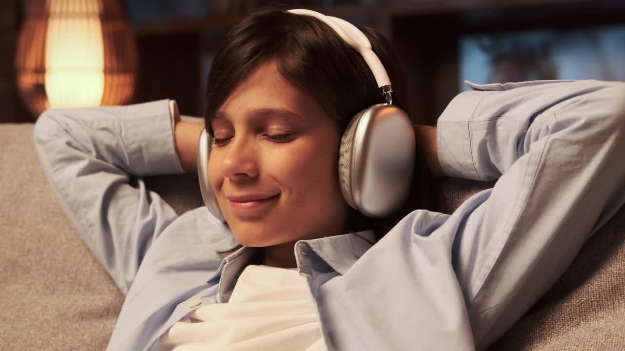 快乐的男生在晚上客厅的沙发上戴着耳机听音乐视频下载