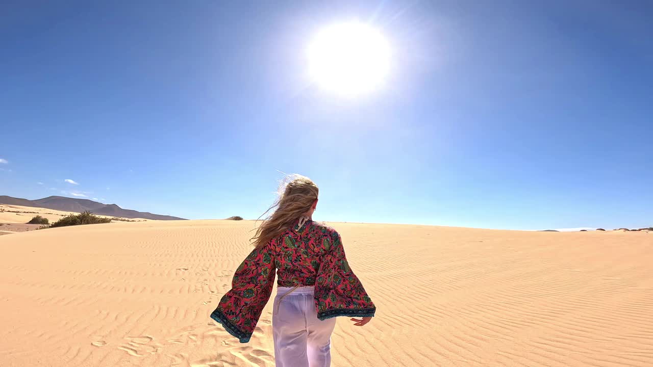 一个快乐的年轻金发女子走在沙丘的沙滩上，享受着假期休闲的乐趣。美丽的女孩发现自由的概念。视频下载