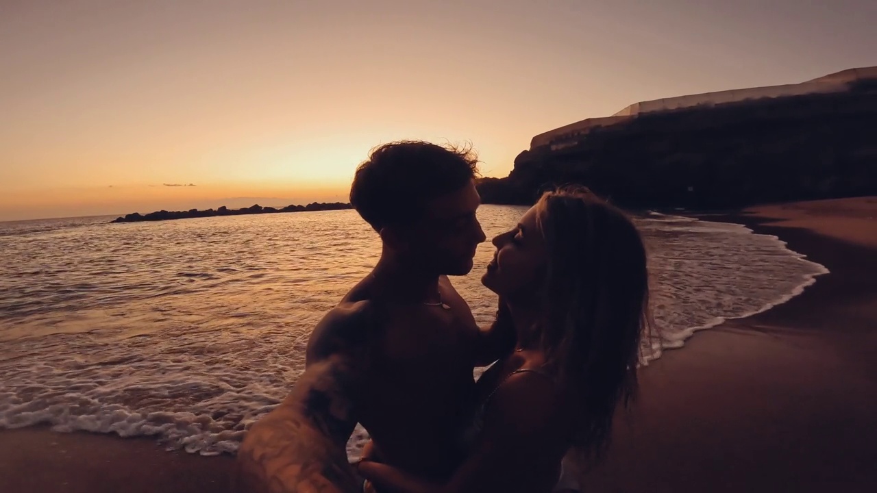 两个年轻的千禧一代在海滩上拥抱亲吻，享受夏日的乐趣，夕阳为背景。度假的浪漫时刻视频下载