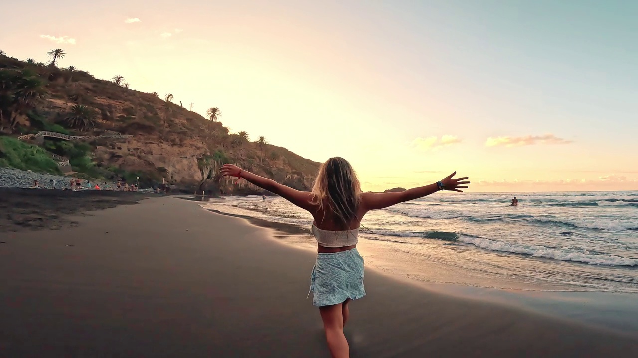 一个快乐的年轻女孩赤脚在沙滩上奔跑和行走，在日落的背景下享受和享受夏天的乐趣。金发碧眼的女人向往着在海边跳舞跳跃的生活方式视频下载