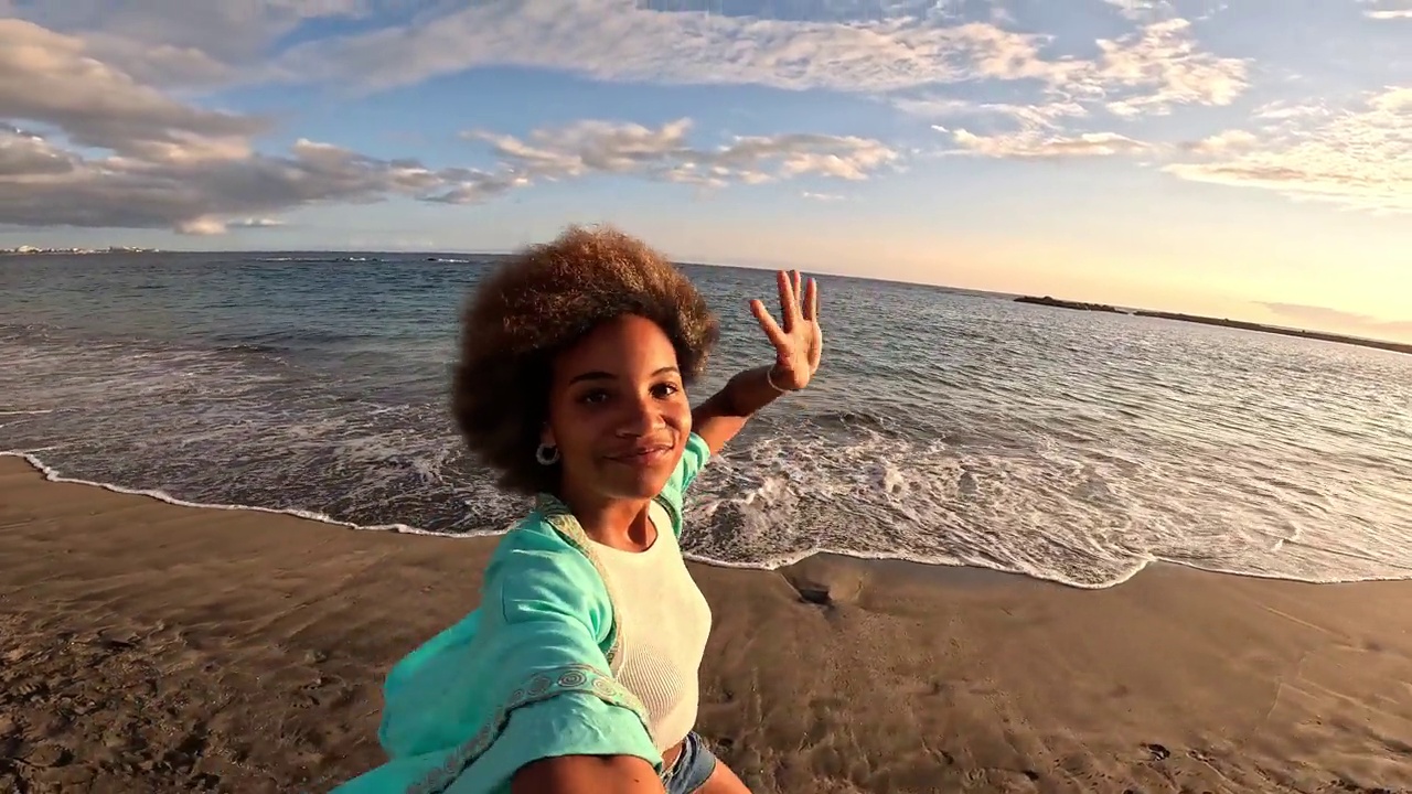一个非洲多种族的年轻美女拿着相机在海滩上自拍，享受夏日户外度假。视频下载