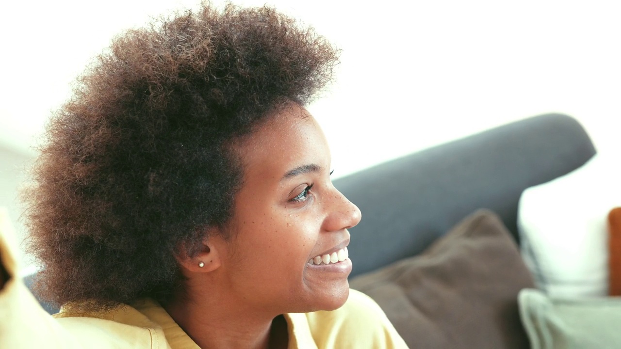幸福的千禧一代黑人混血女孩坐在家里的沙发上，带着牙齿的微笑看着镜头。美丽的非裔美国年轻女子，室内女模特。暴头视频下载