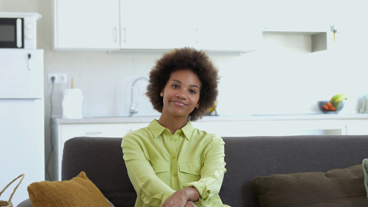 幸福的千禧一代黑人混血女孩坐在家里的沙发上，带着牙齿的微笑看着镜头。美丽的非裔美国年轻女子，室内女模特。暴头视频素材