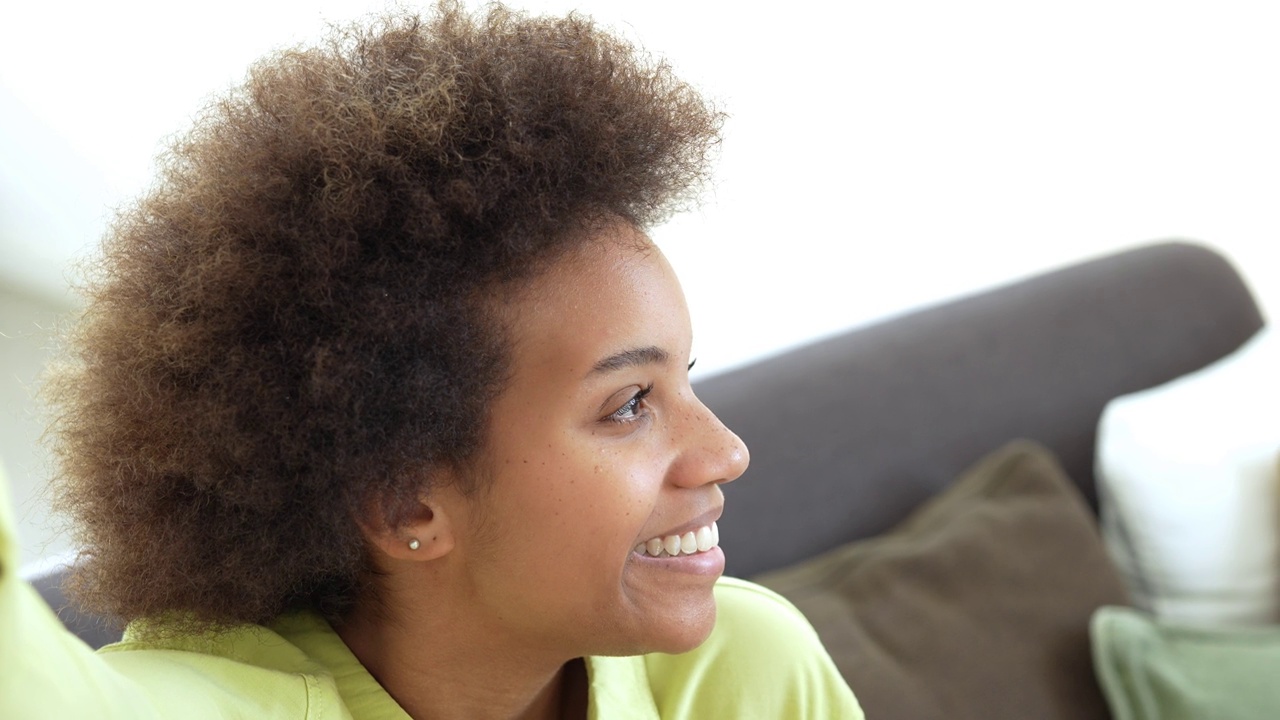 幸福的千禧一代黑人混血女孩坐在家里的沙发上，带着牙齿的微笑看着镜头。美丽的非裔美国年轻女子，室内女模特。暴头视频素材