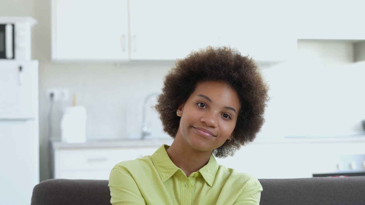 幸福的千禧一代黑人混血女孩坐在家里的沙发上，带着牙齿的微笑看着镜头。美丽的非裔美国年轻女子，室内女模特。暴头视频下载