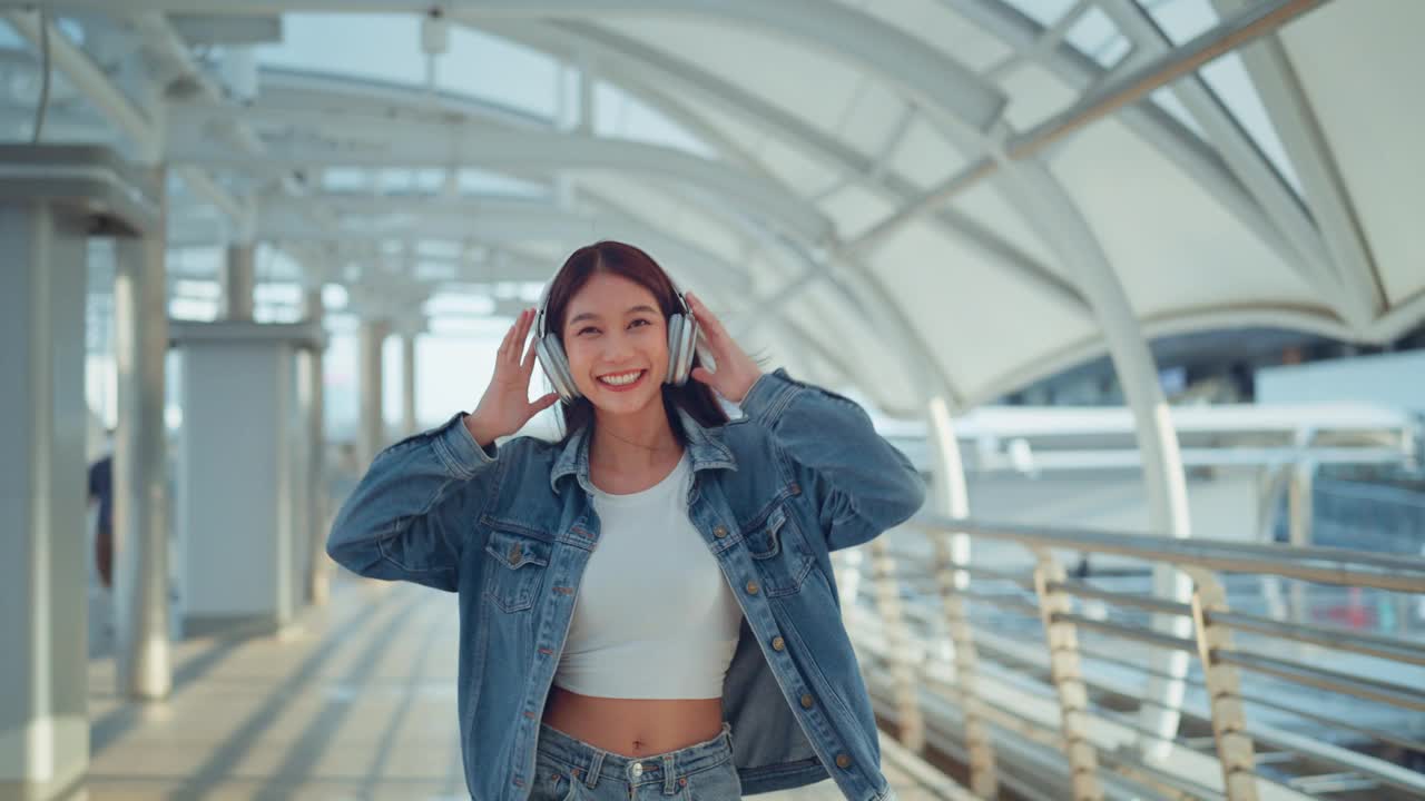 快乐的年轻亚洲女子戴着耳机在的街道上欣赏音乐，微笑的女性独自听音乐跳舞。视频下载