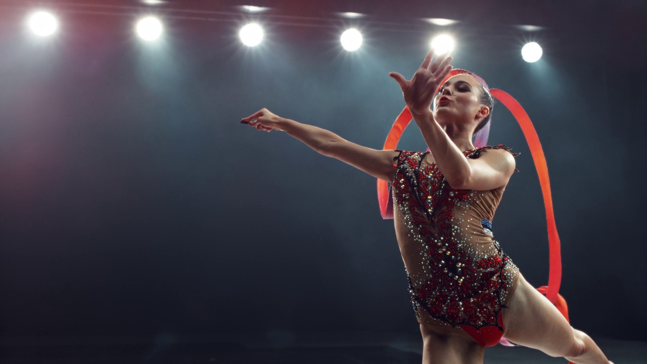 一个现代艺术体操表演的超级慢动作镜头在一个大舞台上的灯光。美丽的女运动员在用红丝带创造形状的同时做出完美的分裂跳跃视频素材