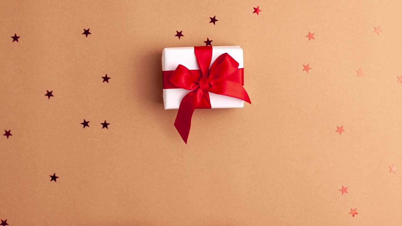 穿着白色毛衣的人的手拿着一个白色的纸礼物，上面有一个红色缎带蝴蝶结，棕色背景上有红色的星星形状。定格动画圣诞假期概念平铺视频下载
