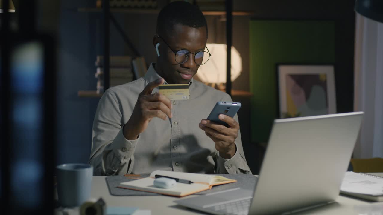 非裔美国人晚上在家用智能手机拿着信用卡网上购物视频素材
