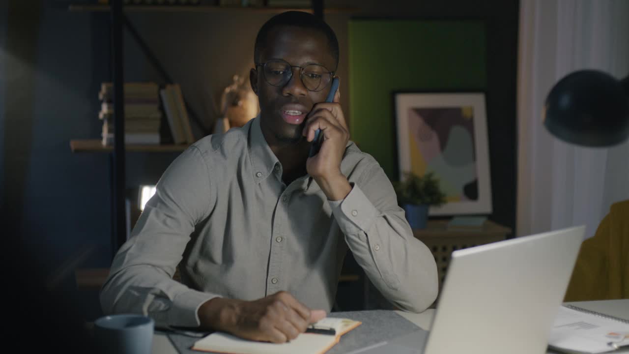 一个非洲裔美国人晚上坐在办公室的办公桌前一边打电话一边喝茶视频素材