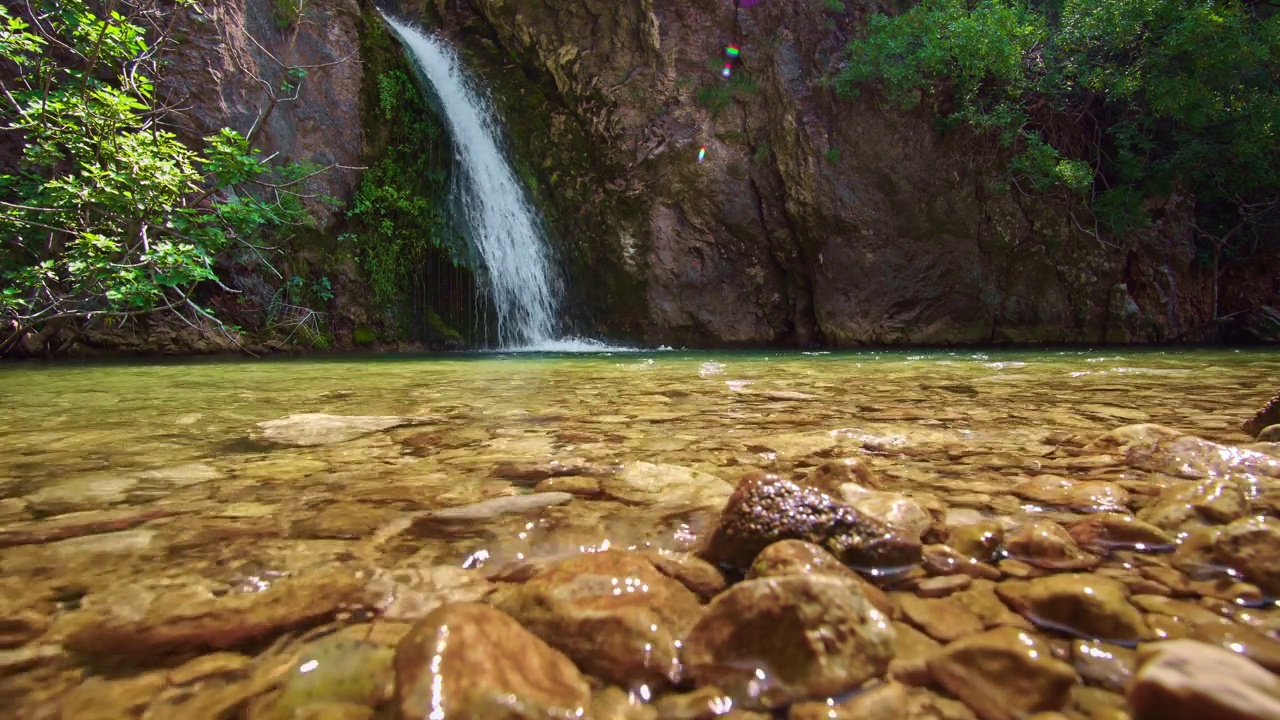 一个瀑布和一个小湖在山上，水倾泻在岩石的背景，一个美丽的夏日景观在一个明媚的阳光灿烂的日子视频下载