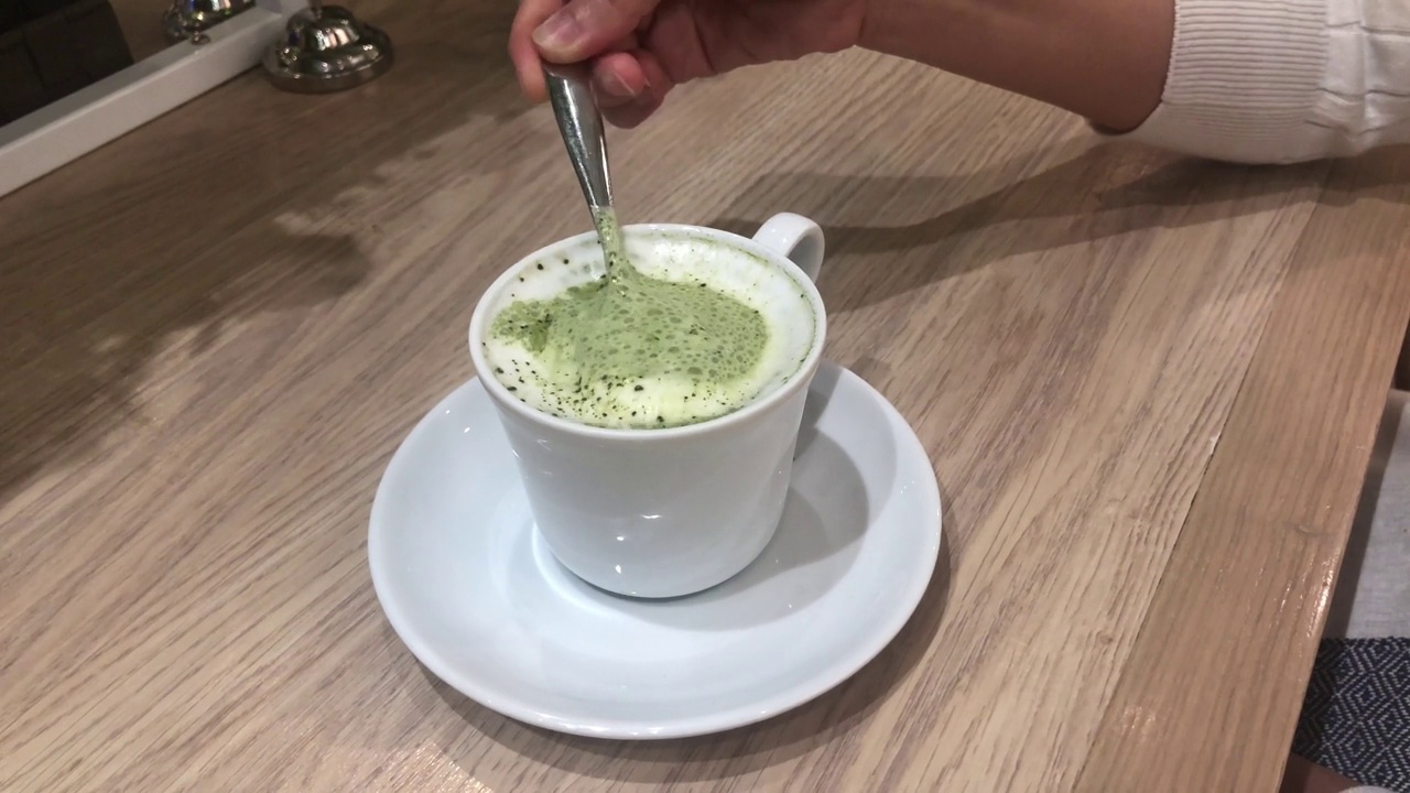 一名亚洲妇女用勺子在蓝色杯子上搅拌日本抹茶绿茶。木桌上健康的绿茶，搅拌着热腾腾的绿茶视频下载