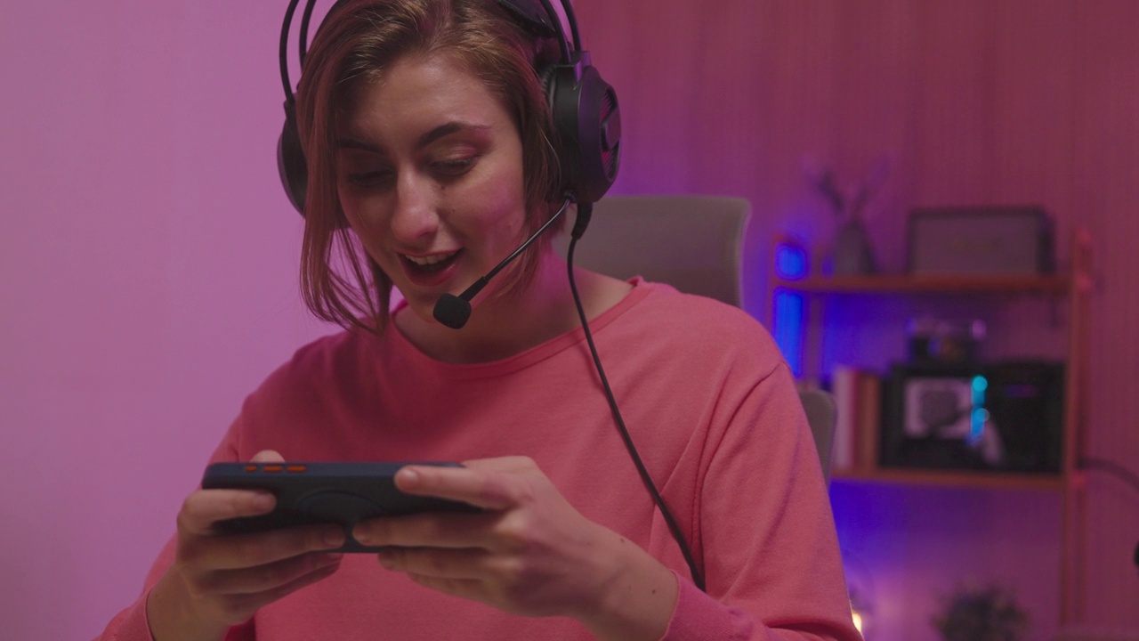 快乐亚洲女孩玩家戴着耳机比赛玩视频游戏在线与智能手机彩色霓虹灯在家里的客厅。视频下载