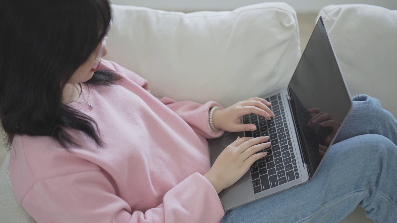 亚洲自由职业女性微笑着躺着，放松地在键盘上打字，在沙发沙发上用笔记本电脑工作。女企业家在客厅里为她的生意工作。商务工作在家的概念。视频下载