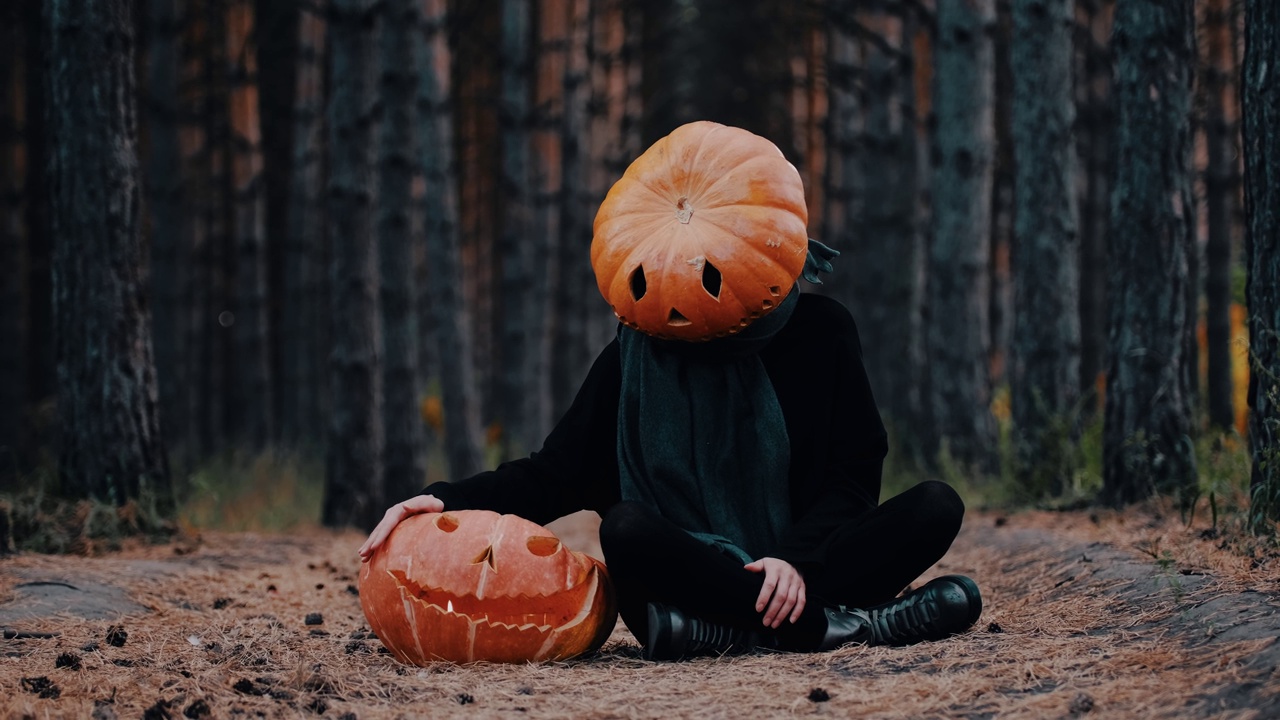 一个穿着黑色衣服，头戴大南瓜头的有趣女孩坐在森林里的地上，旁边是一个大南瓜。万圣节的概念视频下载