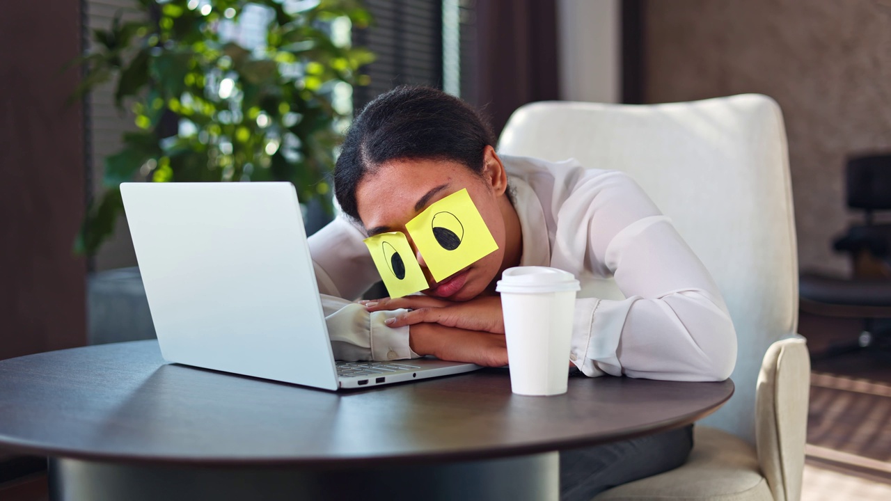 疲惫的非裔美国妇女穿着正装睡在办公桌上，眼睛上贴着黄色贴纸。懒惰的无效率的女性办公室职员靠在打开的无线笔记本电脑旁。视频下载