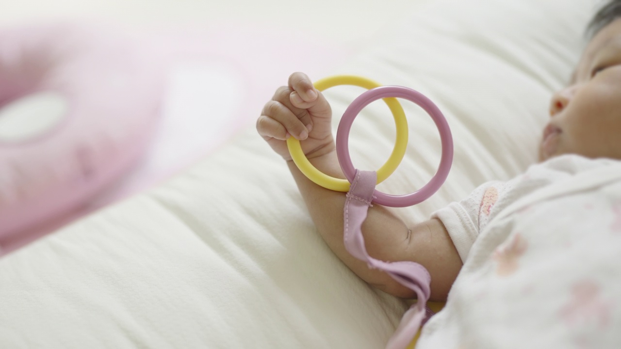 慢镜头:一个刚出生的女婴躺在婴儿床上，抱着并玩着色彩柔和的塑料圈。视频下载