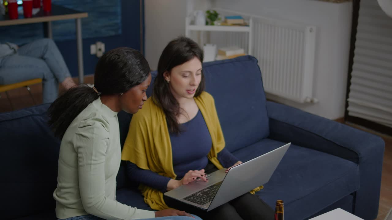 两个多民族的女人坐在沙发上用笔记本电脑看喜剧视频下载