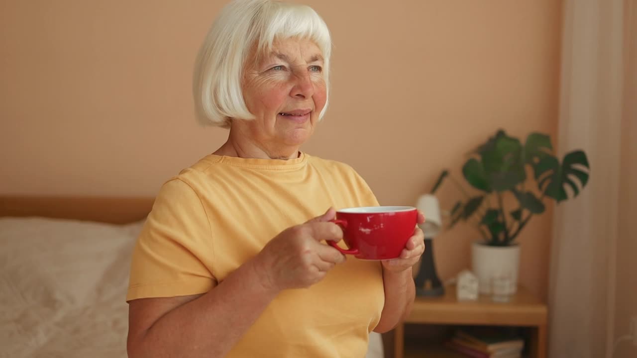 漂亮的60多岁老妇人坐在家里卧室的窗边喝咖啡。视频下载