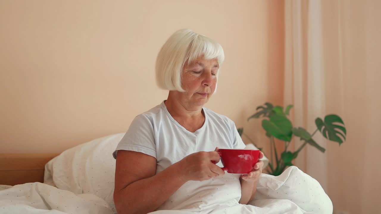 漂亮的60多岁老妇人坐在家里卧室的窗边喝咖啡。视频下载