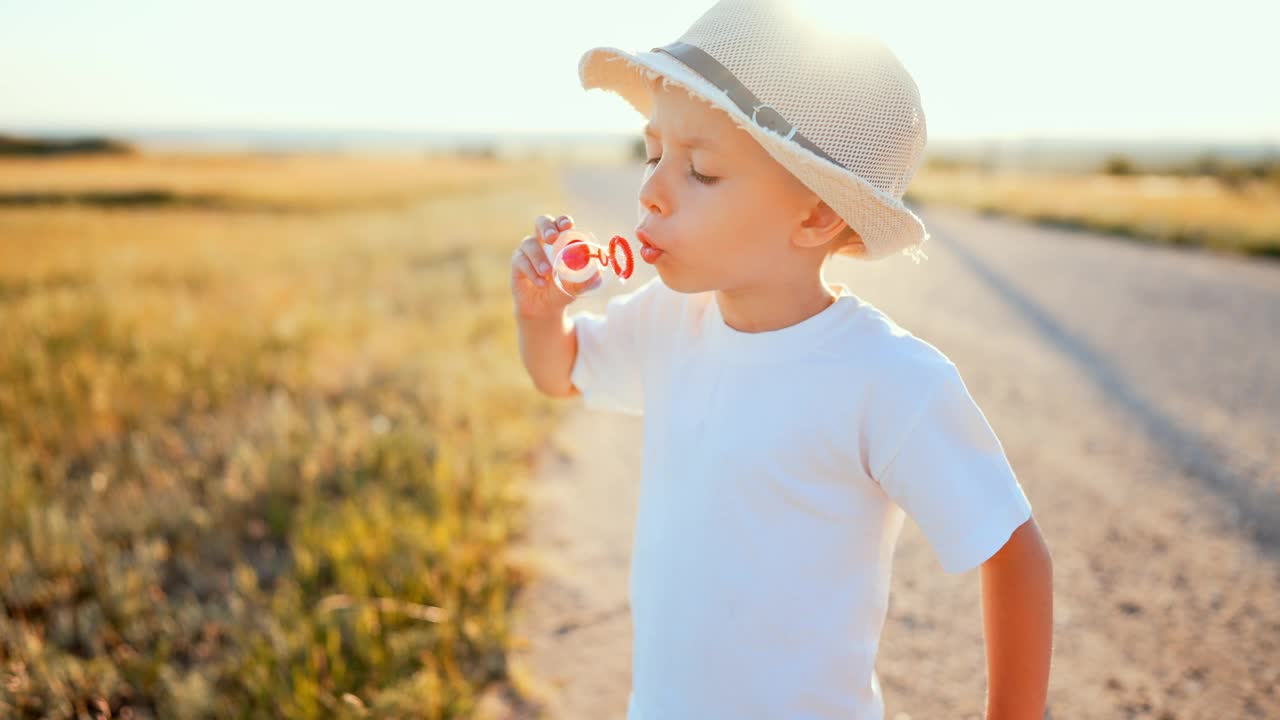 近距离的肖像，男婴戴着草帽站在夏日田野的乡间小路上，在阳光下吹着彩虹反射的肥皂泡。游戏是儿童在户外的休闲活动。视频下载