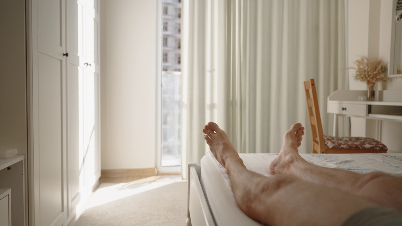 孤独的毛茸茸的男腿在床上扭动脚趾。晴朗温暖的早晨。白色内饰和大全景窗。视频下载