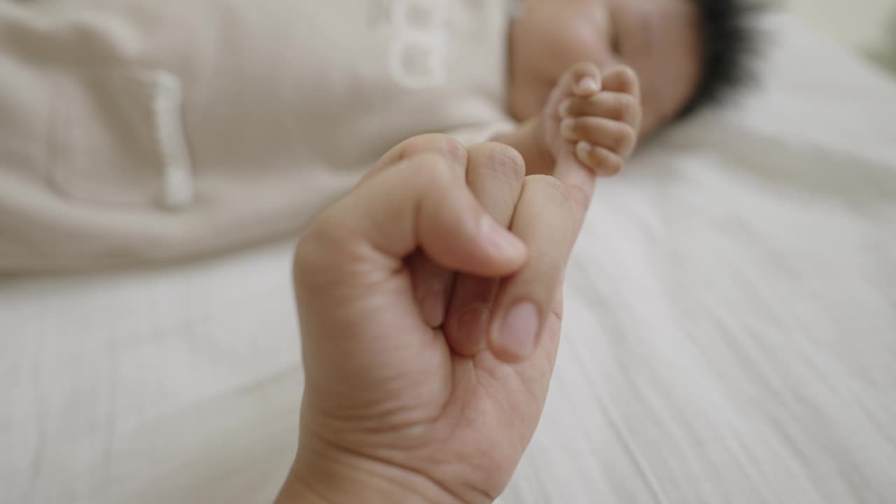 一个刚出生的婴儿在床上打盹时看着并抓着妈妈的手指。视频下载