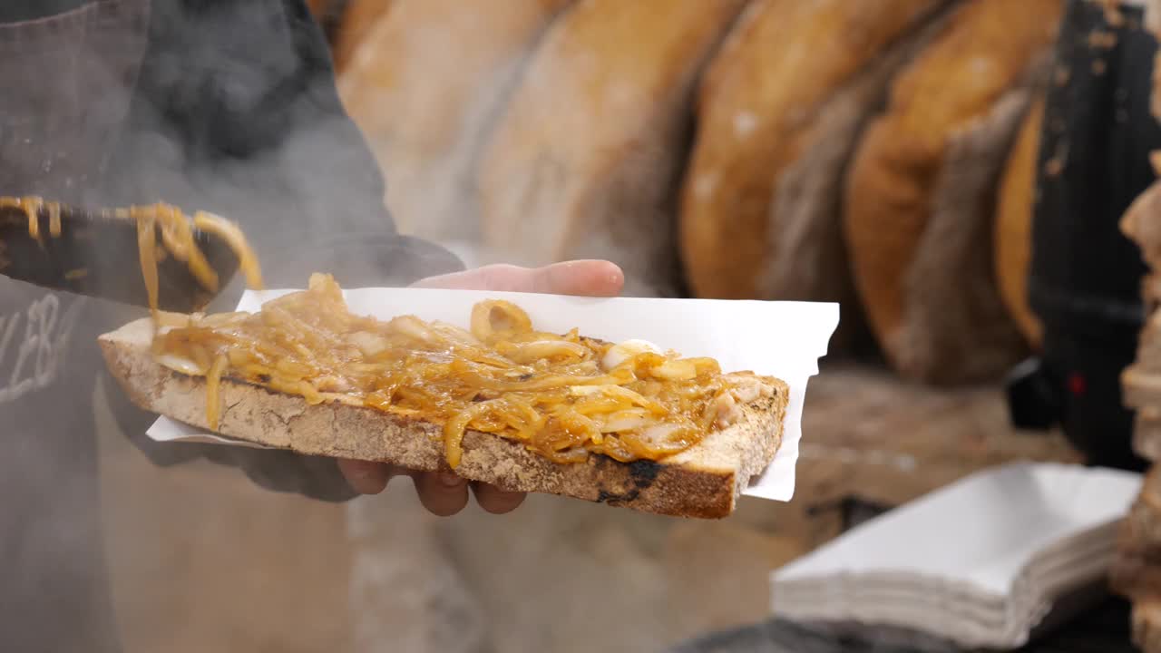 男子将烤好的洋葱铺在一大片面包上，波兰传统美食节市场摊位的特写细节，传统食品的准备，当地街头食品的购买和销售的简单概念视频素材