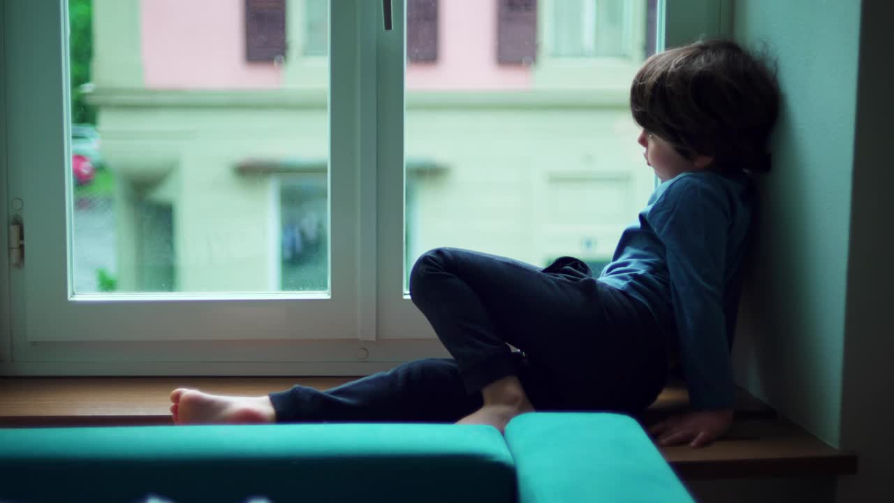 一个无聊的小男孩坐在公寓的窗边，凝视着窗外，感到无聊。孩子在室内感到不安视频素材