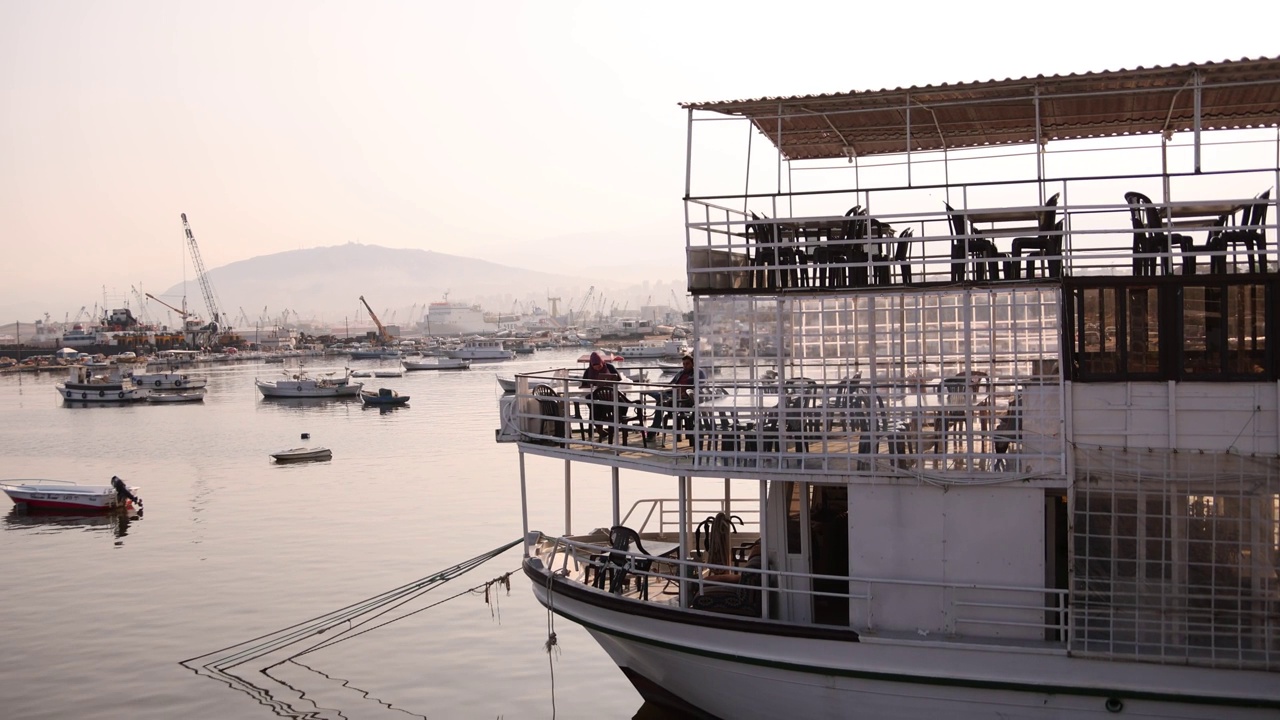 在黎巴嫩北部黎波里的海滨小镇拉米娜，一个古色古香的海港里有一家船上餐厅和咖啡馆，远处是群山视频下载