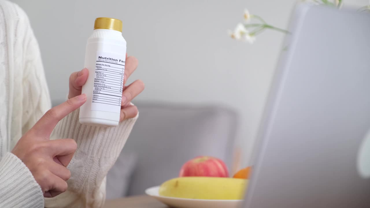 女性营养师通过笔记本电脑向客户展示乳制品瓶上的营养成分标签，为客户提供健康食品建议的特写，制作视频博客，直播流媒体视频下载