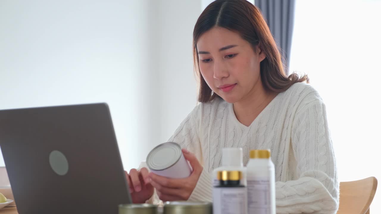 年轻的亚洲女性营养师通过笔记本电脑向客户展示乳制品瓶上的营养成分标签，为客户提供健康食品建议，制作视频博客，直播流媒体视频下载