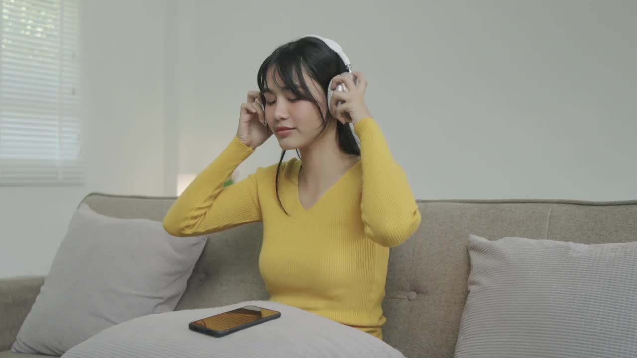 年轻的亚洲女子在家里客厅的沙发上听音乐。快乐的亚洲女性使用手机，戴耳机，坐在沙发上视频下载