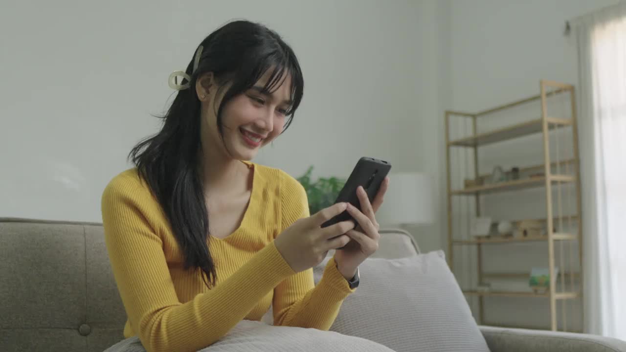 快乐的年轻亚洲女人放松在家里舒适的沙发上发短信在智能手机上，微笑的女孩使用手机，聊天在线消息，从家里网上购物视频下载