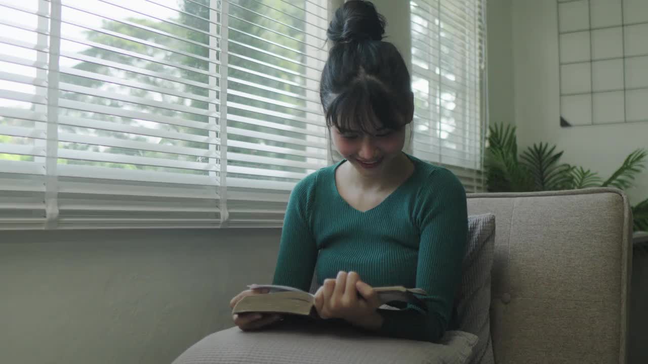 年轻的亚洲女性坐在窗户前的现代沙发上放松在她的客厅看书，喝咖啡或茶在家里视频下载
