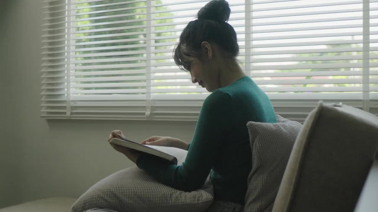 年轻的亚洲女性坐在窗户前的现代沙发上放松在她的客厅看书，喝咖啡或茶在家里视频下载