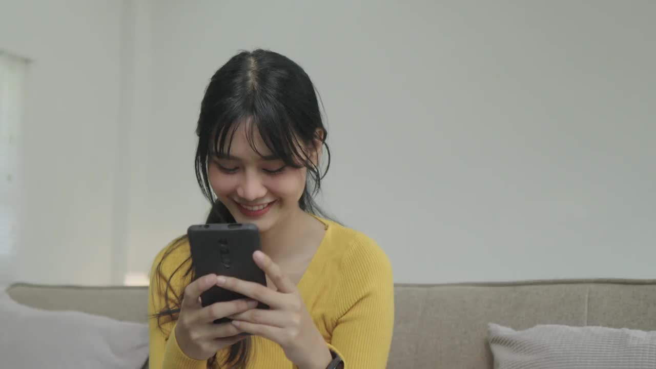 快乐的年轻亚洲女人放松在家里舒适的沙发上发短信在智能手机上，微笑的女孩使用手机，聊天在线消息，从家里网上购物视频下载