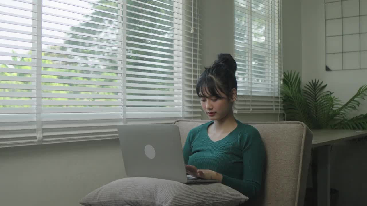 年轻的商业自由亚洲女性工作在笔记本电脑上查看社交媒体，躺在沙发上，放松在家里的客厅。视频下载