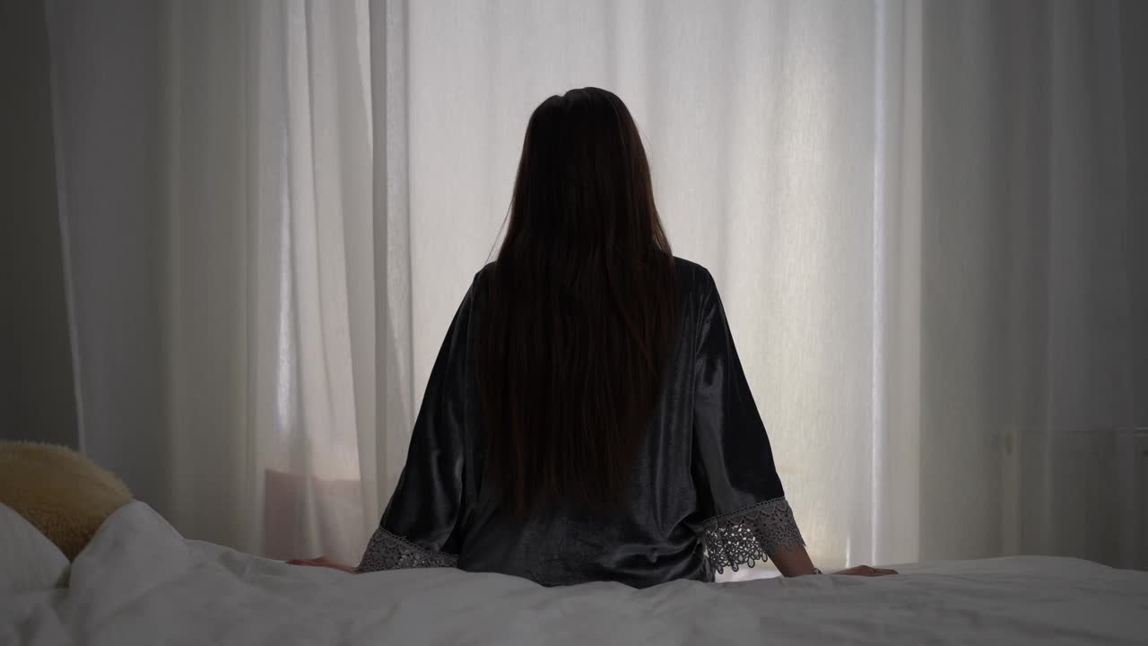 一个穿着睡衣的女人坐在卧室的床上伸展身体，面对着窗帘的窗户。视频下载