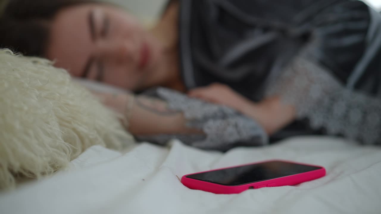 一个穿着漂亮睡衣的女孩躺在柔软的枕头上睡觉。旁边的床上有一个手机，手机开始响了起来。醒来后，女孩手里拿着手机，微笑着。视频下载